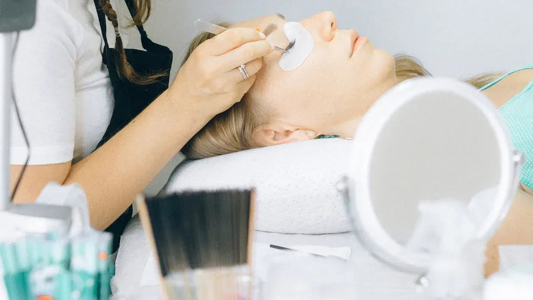 A woman receiving eyelash extensions at a beauty salon, with tools and products nearby.